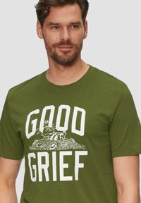 Olijf groene katoenen t-shirt met een grafische print met grote witte tekst "GOOD GRIEF" en een illustratie eronder, ronde hals, korte mouwen.
