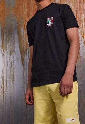 T-shirt en coton noir avec un patch logo sur la poitrine gauche, associé à un short jaune. L'arrière-plan présente un mur peint avec des coulures.