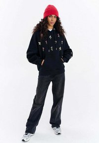 Černá mikina s kapucí s květinovou výšivkou, stažením na šňůrku a přední kapsou. Kombinována s tmavými džíny a červenou pletenou čepicí.