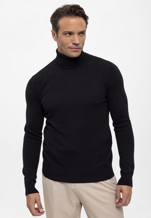 Homme portant un pull noir moulant à col roulé et un pantalon beige, debout et regardant légèrement sur le côté.