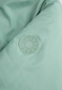 Jasnozielona pikowana tkanina z przezroczystą okrągłą naszywką z wypukłymi literami tworzącymi napis "COCCO DRILL" w formie pierścienia.