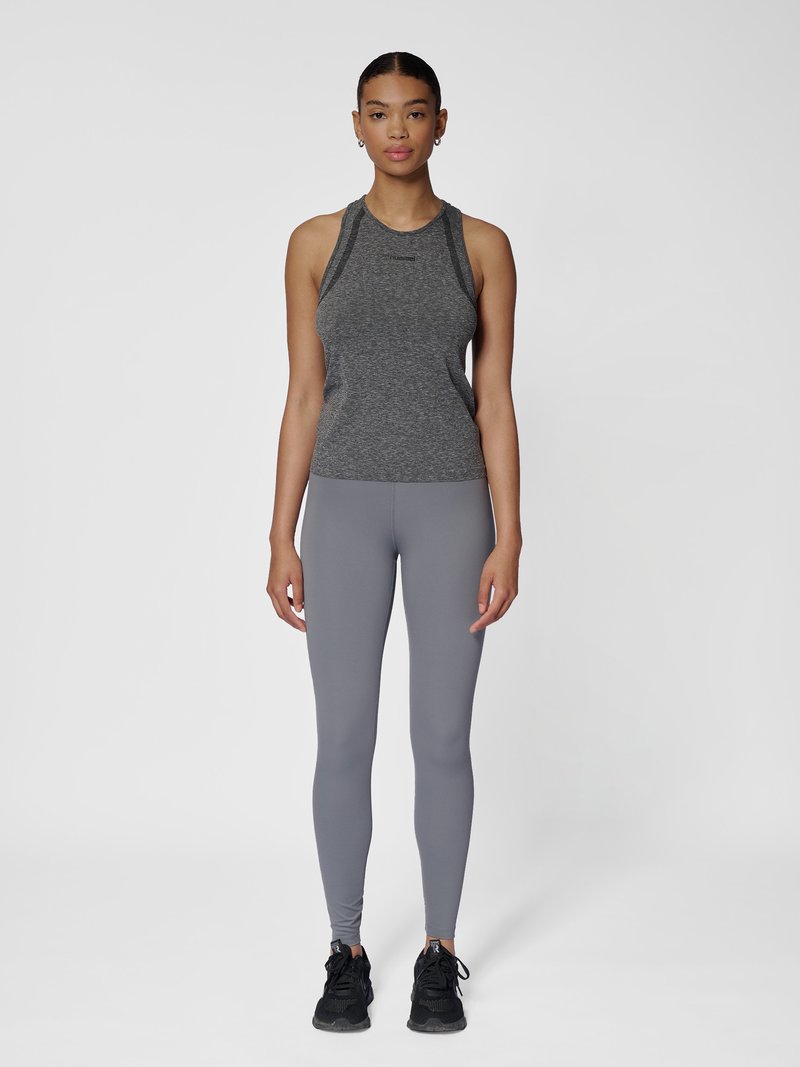 Vrouw met een donkergrijze mouwloze sporttop, lichtgrijze legging en zwarte sportschoenen, staand met het gezicht naar voren op een witte achtergrond.