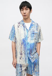 Conjunto de camisa de manga corta y pantalones cortos con un patrón abstracto en azul y blanco, que presenta un cuello con solapa y un bolsillo en el pecho. La tela parece ligera.
