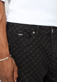 Jean en denim noir avec un motif ton sur ton subtil, des éléments métalliques, des poches avant et une étiquette de marque sur la ceinture.