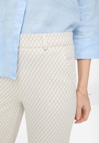 Femme portant une chemise bleu clair et un pantalon beige avec un motif géométrique, montrant sa main droite avec un bracelet fin en argent.