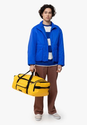 Blue nylon jacket with a collar and pockets, worn over a striped sweater, paired with brown pants. Holding a large yellow duffel bag with black accents.