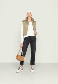 Chaleco acolchado caqui sobre una camiseta blanca de manga larga, combinado con jeans negros y zapatillas blancas. Bolso marrón con detalle de cadena.