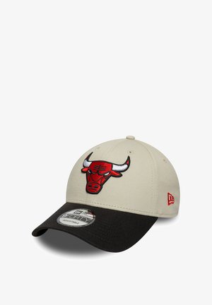 Beige baseball cap with a black brim. Features a red bull logo embroidered on the front. Adjustable strap at the back for fitting.