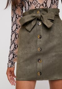 Jupe mini vert olive en tissu doux avec une ceinture large en nœud, comportant six boutons sur le devant et une texture lisse.