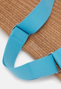 Sac en paille tissée avec un corps texturé de couleur beige, complété par des anses en tissu bleu vif ornées de ferrures argentées et d'un détail de marque embossé.