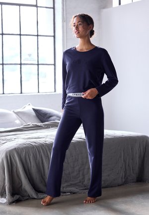 Navy loungewear setti, jossa on pitkähihainen paita, jossa on kohopainettu logo, ja siihen sopivat housut, joissa on valkoinen brändätty vyötärönauha, valmistettu pehmeästä kankaasta.