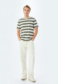 Grøn- og hvidstribet bomulds T-shirt, rund halsudskæring, båret med lyse cremefarvede jeans og beige sneakers. Model står oprejst med hænderne i lommerne.