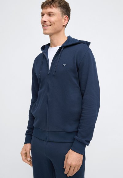 Hoodie zippé bleu marine en tissu texturé, doté d'une capuche avec cordon de serrage, de poignets côtelés et d'un petit logo sur la poitrine. Idéal pour une tenue décontractée.