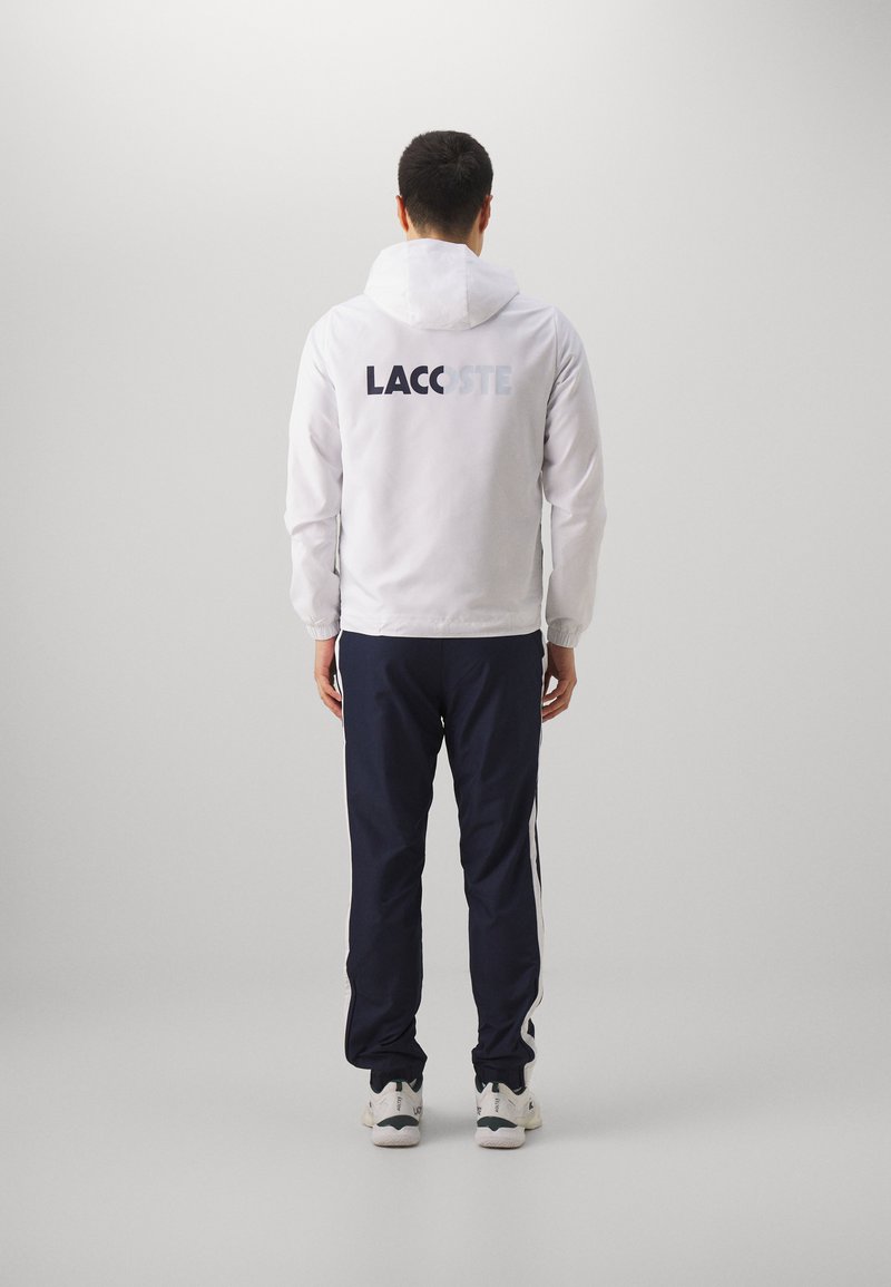 Chaqueta blanca con capucha con el logo "LACOSTE" en la parte posterior, combinada con pantalones de chándal azul marino con rayas blancas a los lados y zapatillas de color claro.