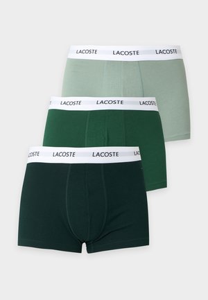 Uomo senza maglietta in piedi a piedi nudi con le braccia incrociate, indossa boxer verde scuro con fascia bianca etichettata "Lacoste", su sfondo bianco semplice.