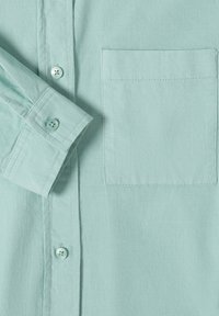 Chemise en velours côtelé turquoise clair avec poche poitrine avant, boutons visibles et revers plié sur la manche gauche.