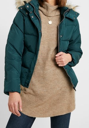 Kvinde iført en teal puffjakke med hætte foret med pels over en beige strikket sweaterkjole og mørke jeans, som holder jakken åben ved lynlåsen.
