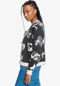 Black knitted cardigan featuring white abstract patterns, pink ribbed cuffs, and collar. Model styled with denim overalls and braided hair.