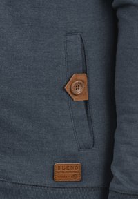 Marineblauwe hoodie met een gestructureerde stof, een bruin knoopaccent op de zak en een leren label met reliëftekst.