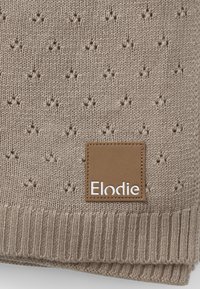 Beige neulotut viltti, jossa on tekstuuri ja ribbääre. Mukana on suorakulmainen ruskea nahkaleima, jossa lukee "Elodie" kohokuviona.