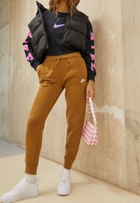 Svart pufferväst, mörkblå sweatshirt med rosa detaljer, bruna joggers, vita sneakers och en rosa rutmönstrad handväska.