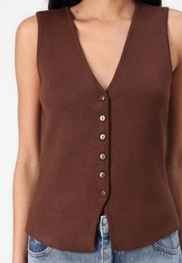 Gilet en tricot sans manches de couleur marron avec un décolleté en V, doté d'une fermeture à boutons et d'un ourlet légèrement fendu, fabriqué à partir d'un matériau texturé doux.