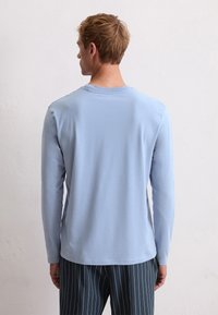 Camisa de manga larga azul claro hecha de una tela suave, con un cuello redondo y un corte relajado. Costuras visibles y dobladillo ligeramente curvado.