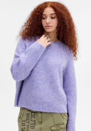 Jeune femme aux cheveux roux bouclés portant un pull violet doux et un pantalon vert olive avec un texte noir, regardant vers le bas avec la main près du menton.