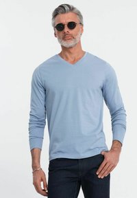 Camicia a maniche lunghe di colore blu chiaro con scollo a V realizzata in tessuto morbido, abbinata a jeans in denim scuro. Modello che indossa occhiali da sole neri a forma rotonda.