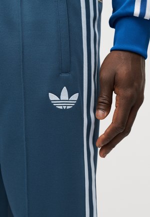 Dark-skinned hand next to blue Adidas track pants with white three-stripe detailing and white trefoil logo near pocket.