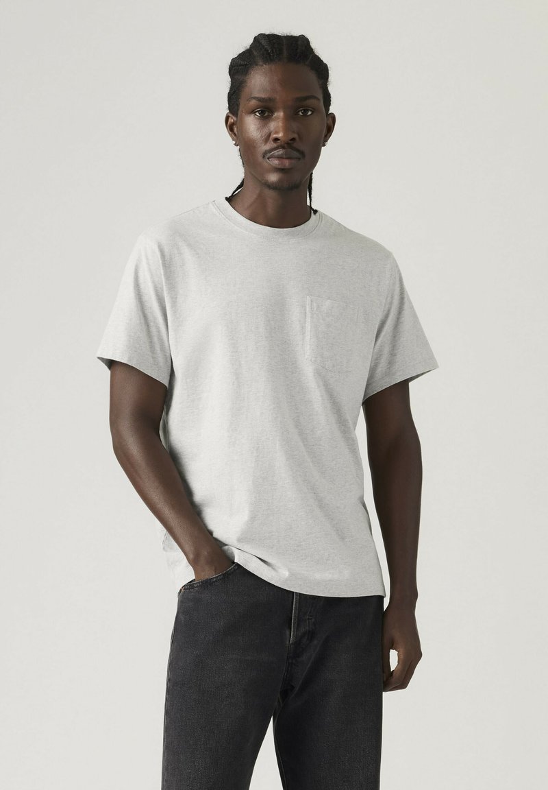 Mann mit geflochtenem Haar, der ein hellgraues T-Shirt mit Tasche und schwarze Jeans trägt, steht mit einer Hand in der Tasche vor schlichter Kulisse.