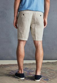 Bărbat îmbrăcat în pantaloni scurți bej, cămașă albastră și pantofi slip-on negri, stând pe un covor cu model, în fața unui perete gri.