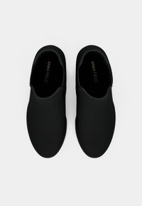 Un par de zapatos negros tipo slip-on con puntas redondeadas y lados elásticos, vistos desde arriba sobre un fondo blanco.