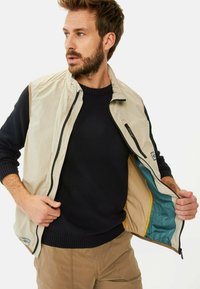 Beige leichte Weste mit Reißverschluss, kontrastierenden braunen Akzenten und einem Innenfutter mit türkisfarbenen und gelben Abschnitten. Über einem dunklen Pullover getragen.