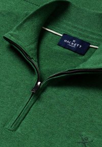Zelený pletený sveter s polozipsom, textúrovaným golierom, vyšívaným značkovým logom blízko lemu a tmavomodrou štítkom Hackett London vo vnútri goliera.