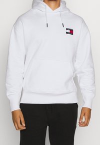 Person wearing white hoodie with front pocket and small Tommy Jeans logo on chest, paired with black pants, standing against plain background.