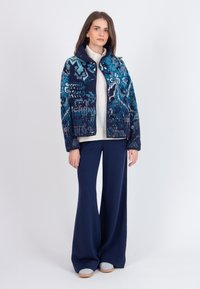 Jūras zils rakstains žakets ar zilo un balto akcentu, pieci pogas, adīts tekstūras; kombinēts ar pieguļošām jūras zilām biksēm un gaišām kurpēm.