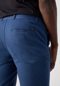 Pantalones de algodón azul marino con una textura suave. Presentan un bolsillo trasero, detalles de costura y una etiqueta de la marca en la parte trasera.