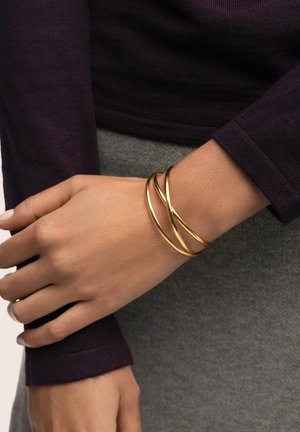 Mano que lleva una pulsera de brazalete dorada retorcida sobre un top de manga larga color púrpura oscuro y fondo de falda o pantalones grises.