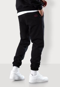 Schwarze Sweatpants mit elastischen Bündchen, die ein kleines pinkes Logo auf der Gesäßtasche haben, getragen mit weißen Sneakern und hochgerollten weißen Socken.