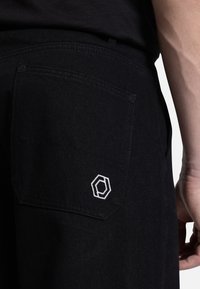 Black denim pants with a back pocket featuring white geometric embroidery. Smooth texture and tailored fit detail visible.