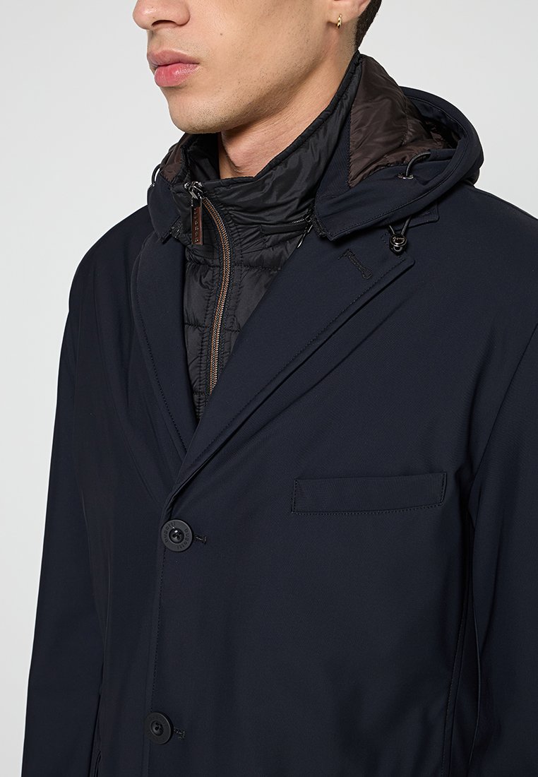 Marineblauer Mantel mit abnehmbarer Puffer-Fütterung, Reißverschlusskragen, zwei Knöpfen und einer Brusttasche. Glattes Material mit taillierter Passform.