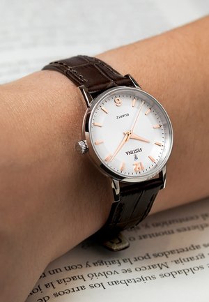 Wrist with a Festina quartz watch showing 3:37, brown leather strap, resting on an open book with black text on white pages.