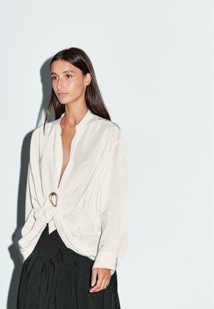 Woman with long dark hair wearing a white long-sleeve blouse with a gold buckle and a black pleated skirt against a plain white background.