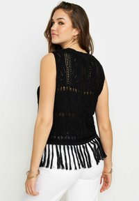Top nero lavorato a maglia con orlo frangiato, caratterizzato da un motivo testurizzato e design senza maniche. Indossato con pantaloni bianchi.