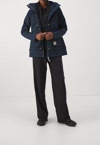 Navy waterproof jacket with a hood, zip front, two large front pockets, and a logo patch. Paired with black trousers and sturdy footwear.