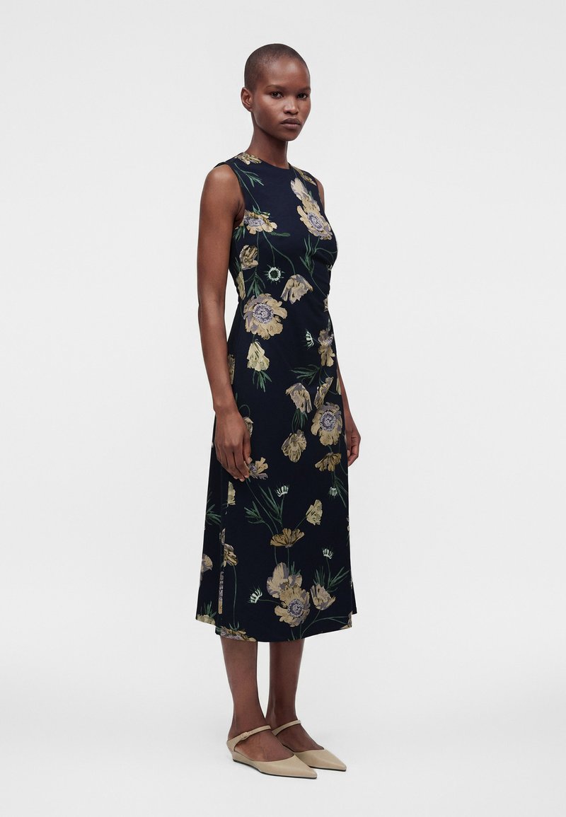 Woman stands facing left, wearing a sleeveless dark floral midi dress and beige pointed flats against a plain light background.