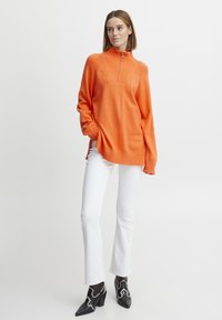 Pullover orange en tricot à col montant avec zip quart, présentant une texture côtelée et des fentes sur les côtés, associé à un jean droit blanc et des talons noirs.