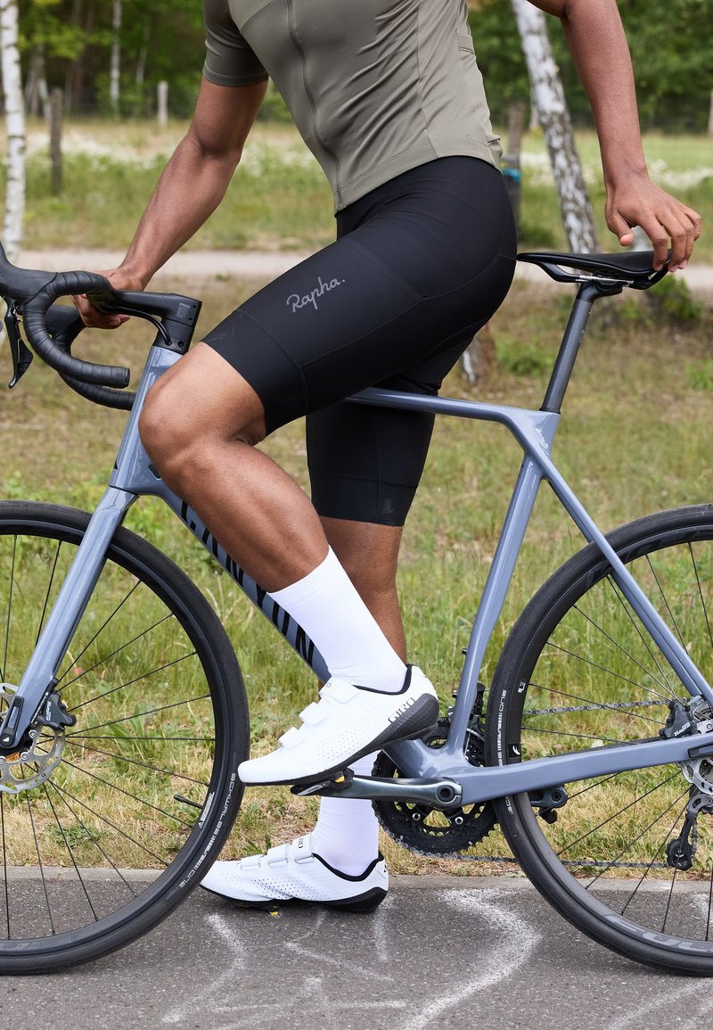 Cycliste sur un vélo gris portant un maillot olive ajusté, un short noir, des chaussettes blanches et des chaussures blanches à bandes Velcro, sur un chemin pavé.