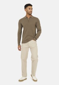 Pullover khaki con collo a zip, maniche lunghe e texture a coste, abbinato a pantaloni beige chiaro e sneakers bianche.